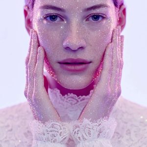 A close-up portrait of a woman with freckles, her face and hands shimmering with thousands of tiny, glowing pink and gold light particles.